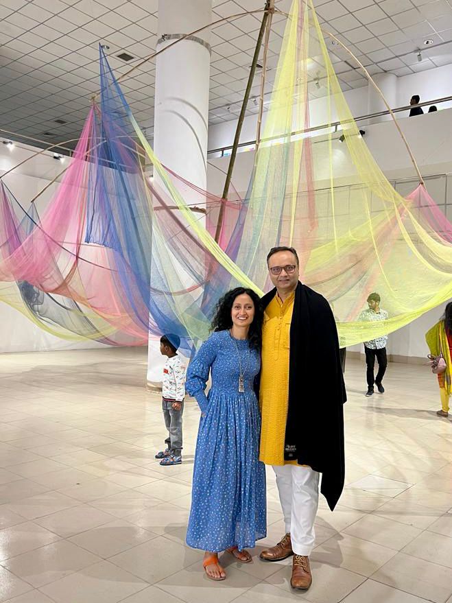 A man in a yellow dress and white trousers wearing a black cardigan standing with a woman in blue dress in front of a multicoloured net hanging from the ceiling