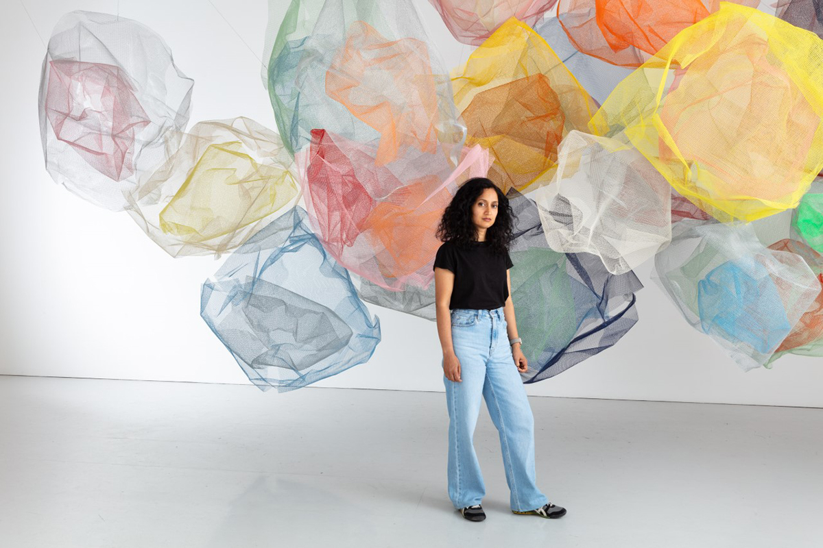 A woman wearing jeans and a black t shirt standing in front of multicoloured nets hanging on a wall