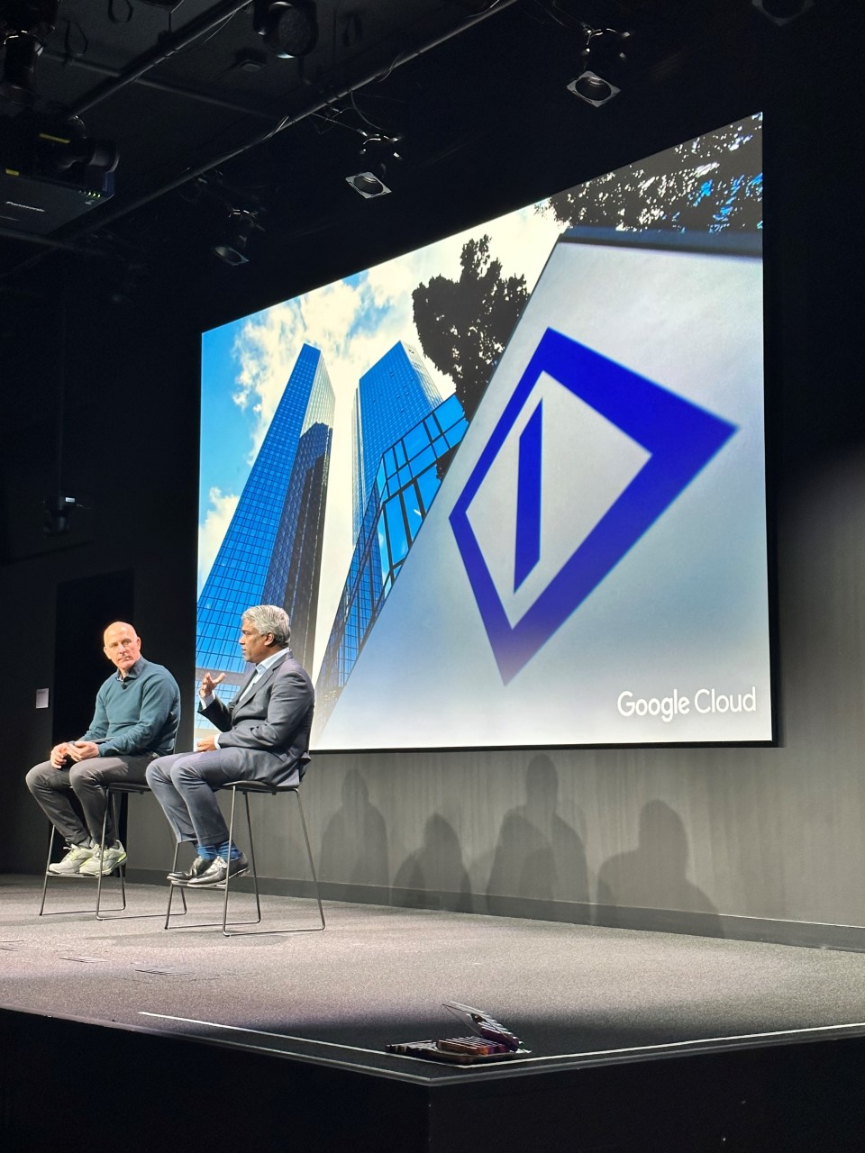 Two men sitting on stools on a stage with a Deutsche bank logo on a screen behind them