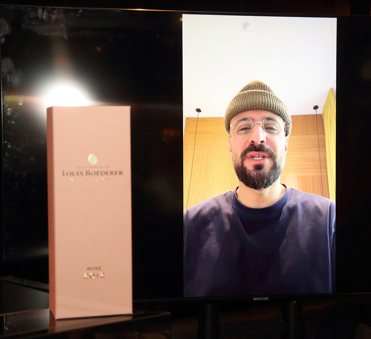 A man wearing a hat with a beard on a screen next to a pink case of champagne