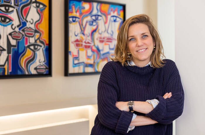 A woman wearing a blue jumper with her arms folded standing in front of colourful paintings