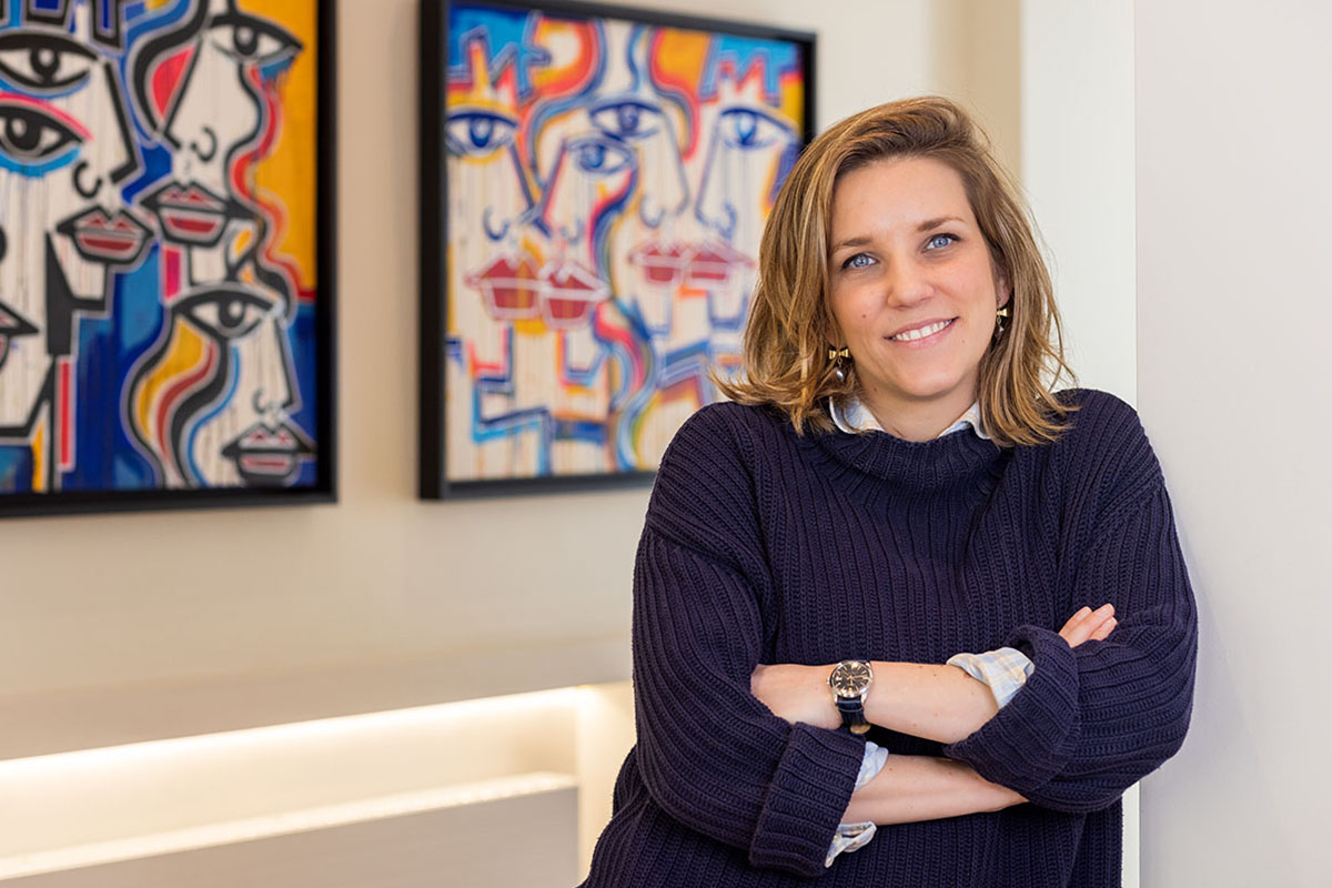 A woman wearing a blue jumper with her arms folded standing in front of colourful paintings