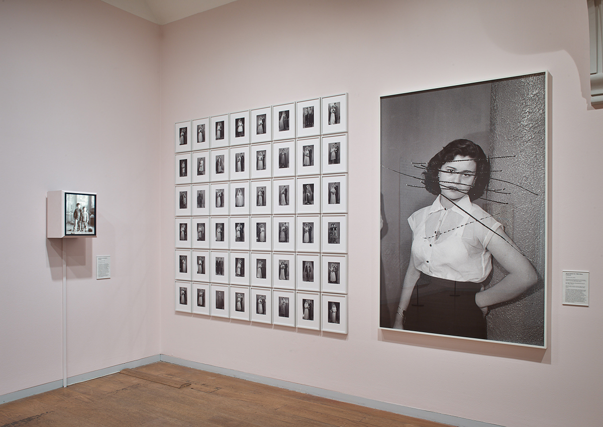 A black and white photograph of a woman in a shirt and black skirt next to lots of small photographs on a gallery wall