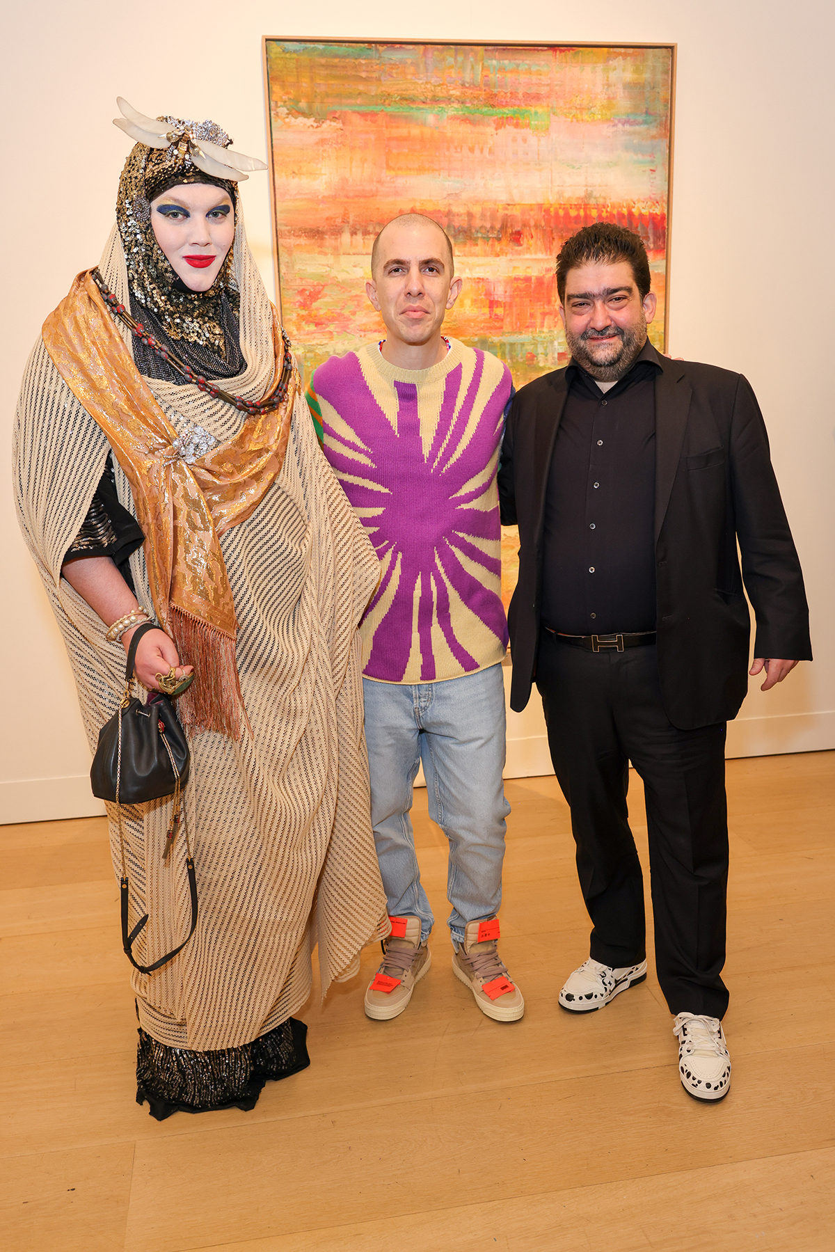 A man in a black suit standing next to a man wearing a purple tie dye jumper, blue jeans and orange trainers standing next to a man wearing a beaded dress and head cover