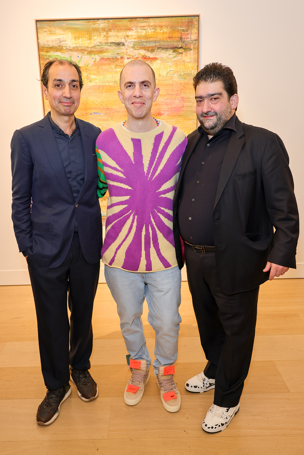 a man wearing a purple tie dye jumper, blue jeans and orange trainers standing between two men in dark suits