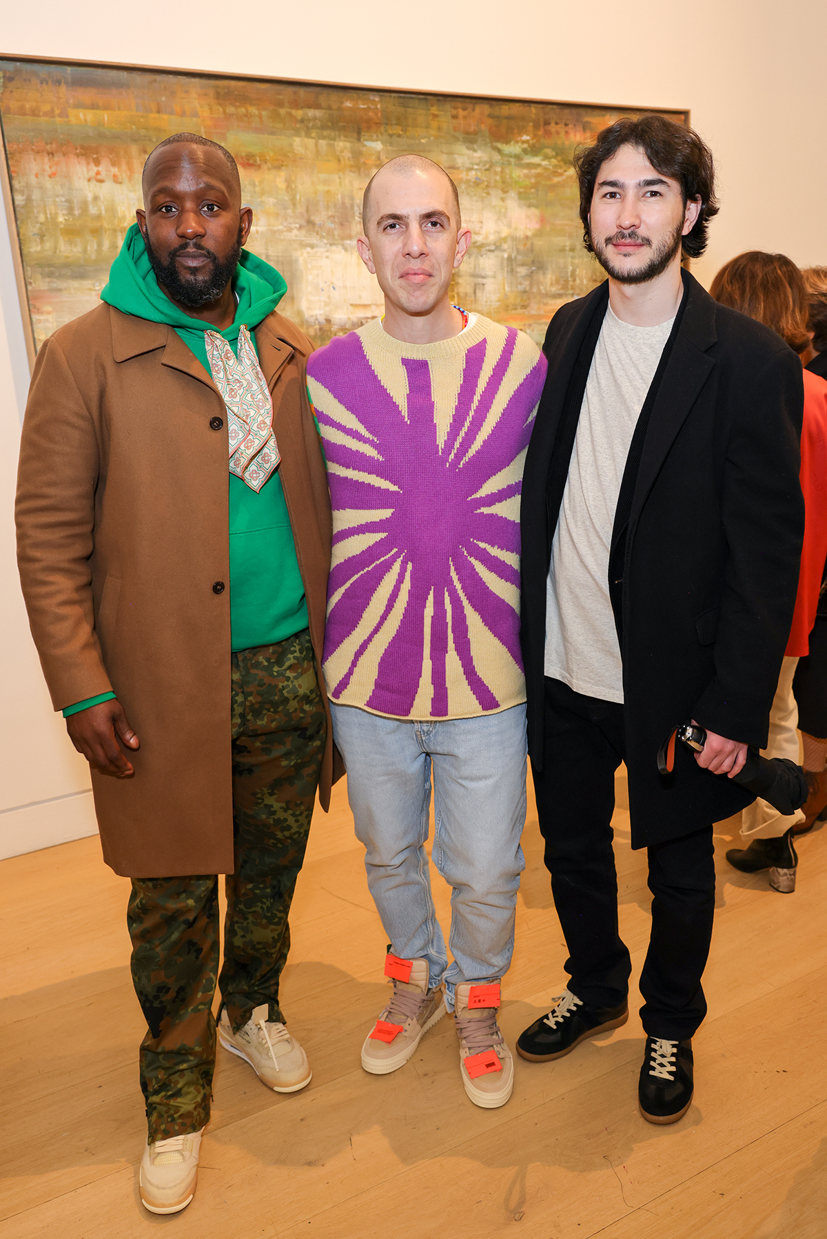 a man wearing a purple tie dye jumper, blue jeans and orange trainers standing next to a man wearing a green hoodie and brown and on the side a man in a black jacket and jeans and white top