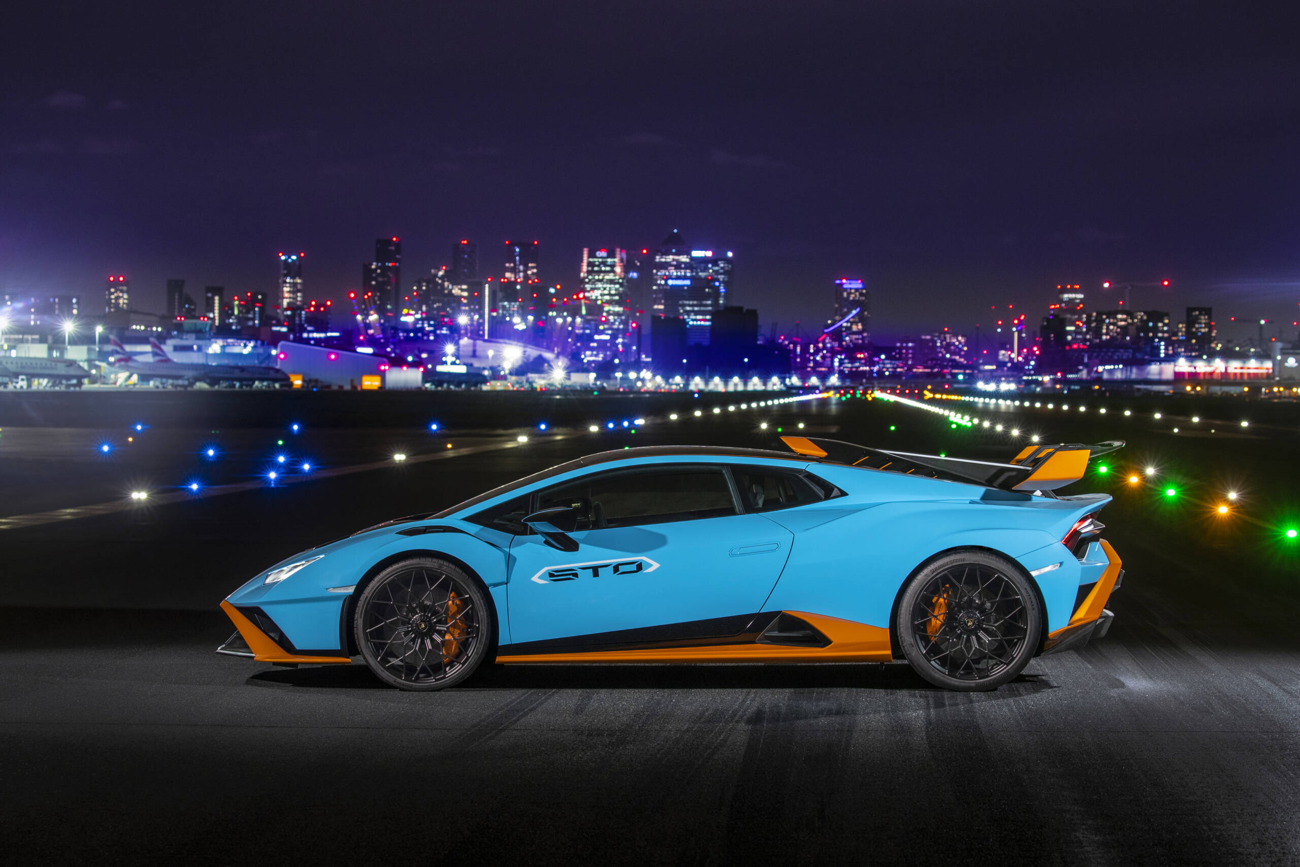 A blue and orange Lamborghini on a road at night with a lit up skyline behind it