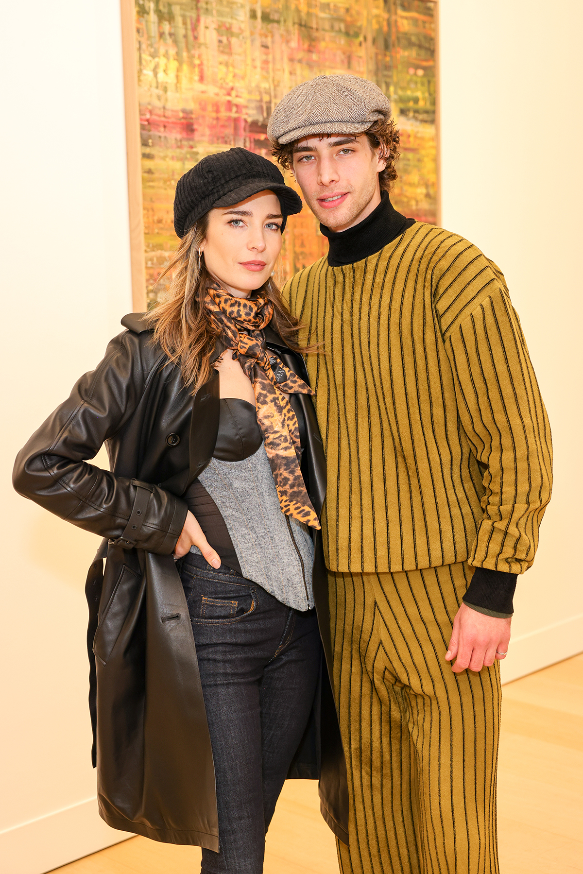 A man wearing a yellow and black striped coord standing next to a woman wearing a black hat, jacket and jeans with a grey striped top