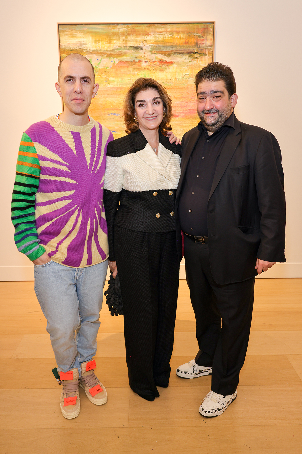 A woman in a black and white suit standing between a man in an all black suit and another man wearing a purple tie dye jumper, blue jeans and orange trainers