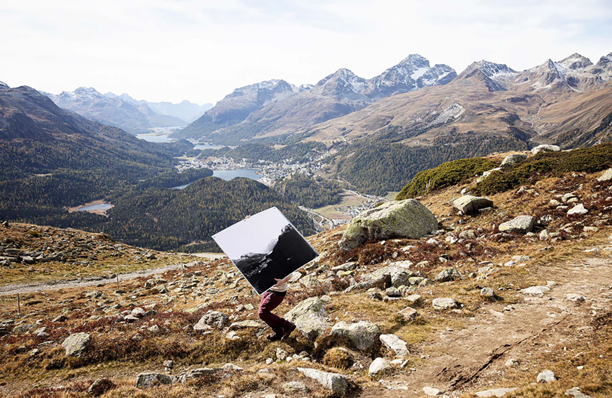 A man holding a mirror on a dry mountain with a town in the dsisance