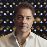 A man wearing a white shirt and necklace standing in front of bottles of wine on shelves