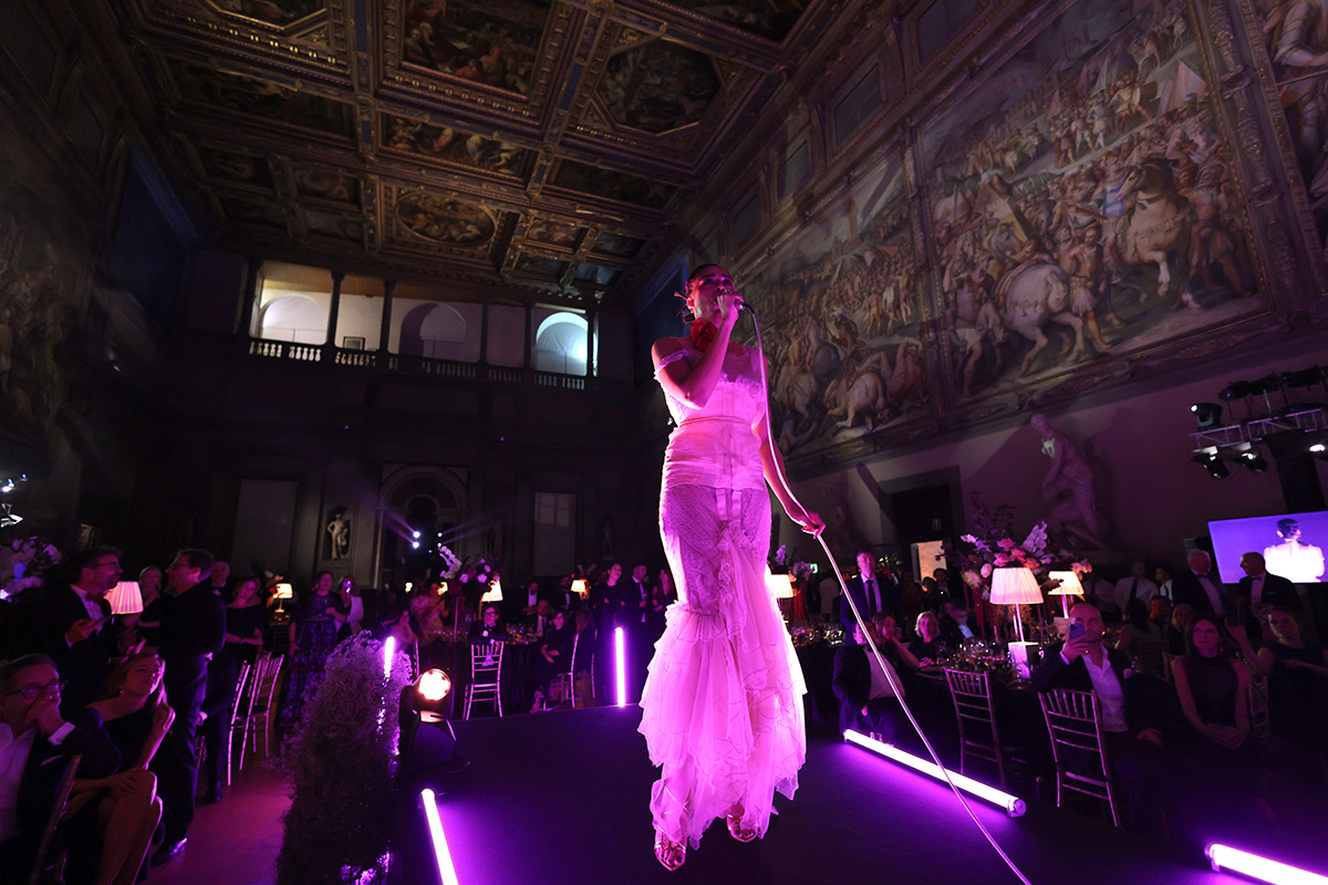 A woman in a pink dress singing on a stage whilst people sit at tables around the stage