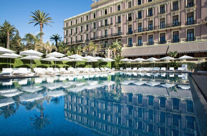 A swimming pool surrounded by white umbrellas and deck chairs with a hotel in the background