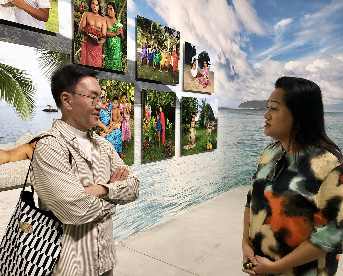 Two people speaking to each other by a wall with a picture of a beach and paintings on top of it