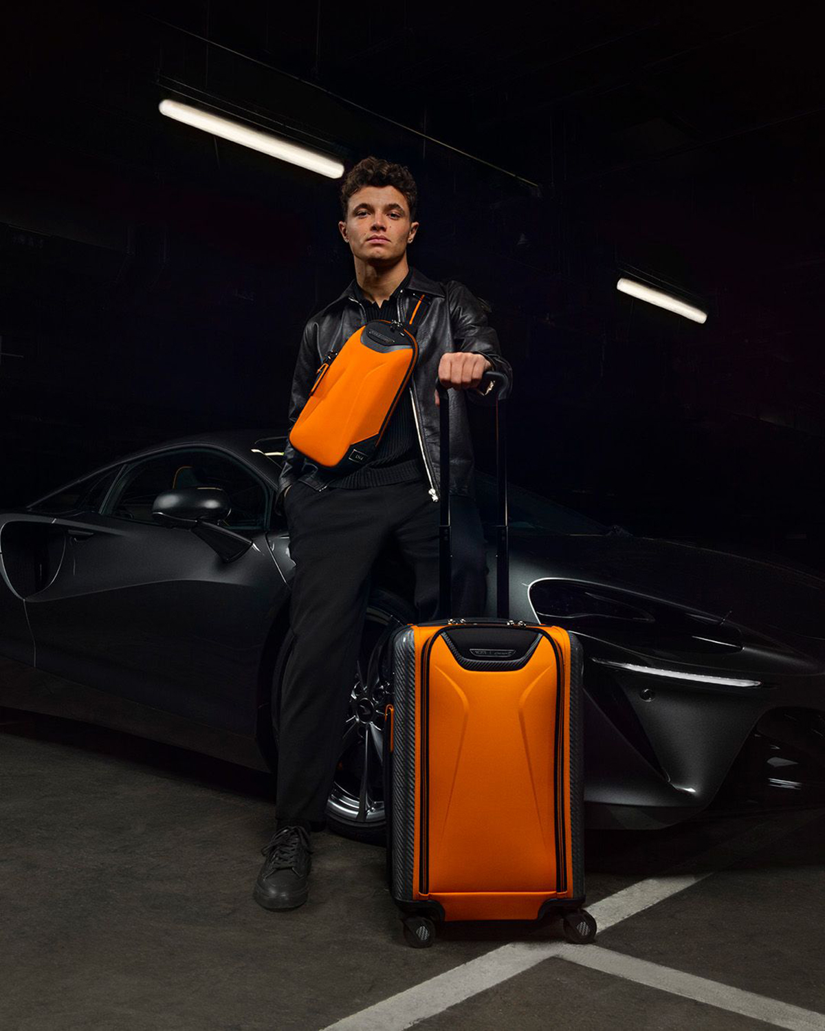 Lando Norris holding an orange rucksack and standing next to an orange suitacase