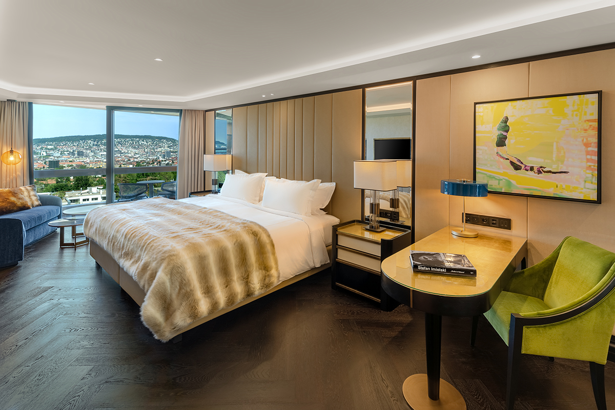 A bedroom with a view of a city and beige headboard and throw on the bed