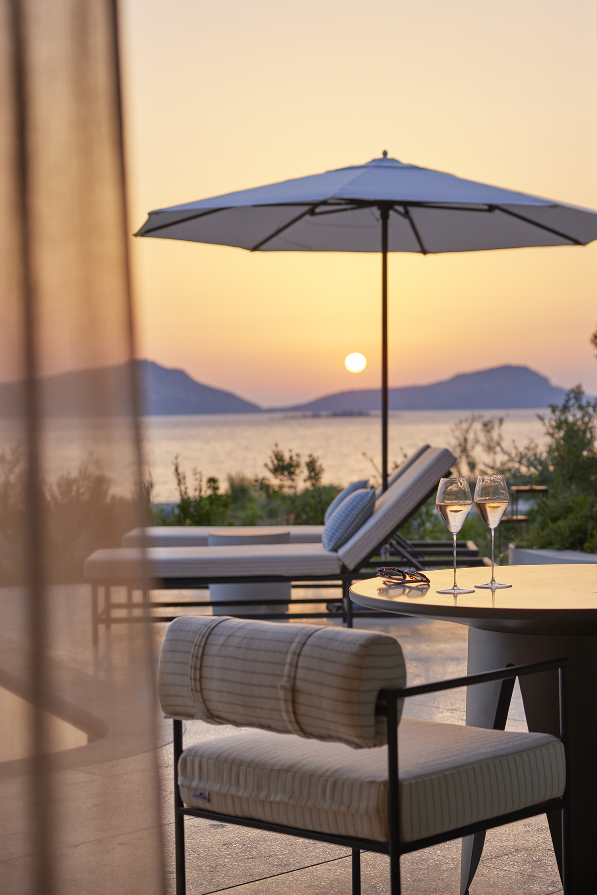 A deck chair and parasol with mountains and see in the distance with a sunset