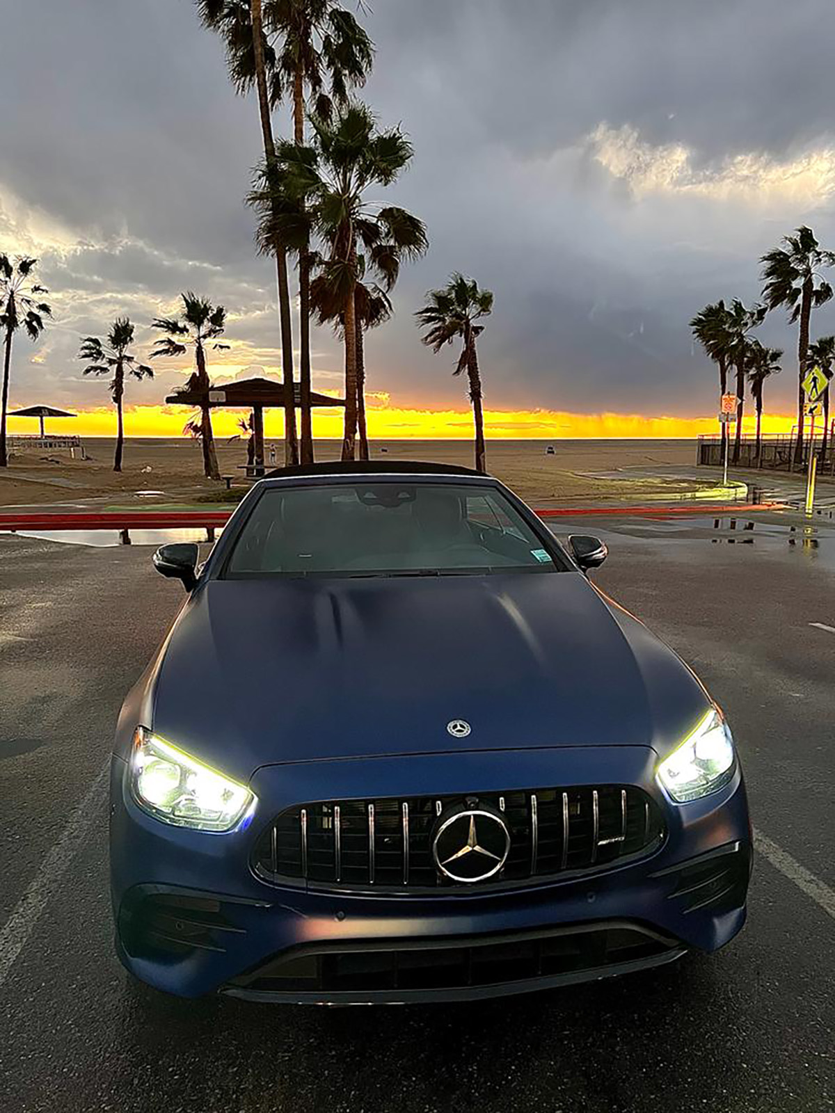 A blue Mercedes with its headlights on with a sunset and palm trees behind it