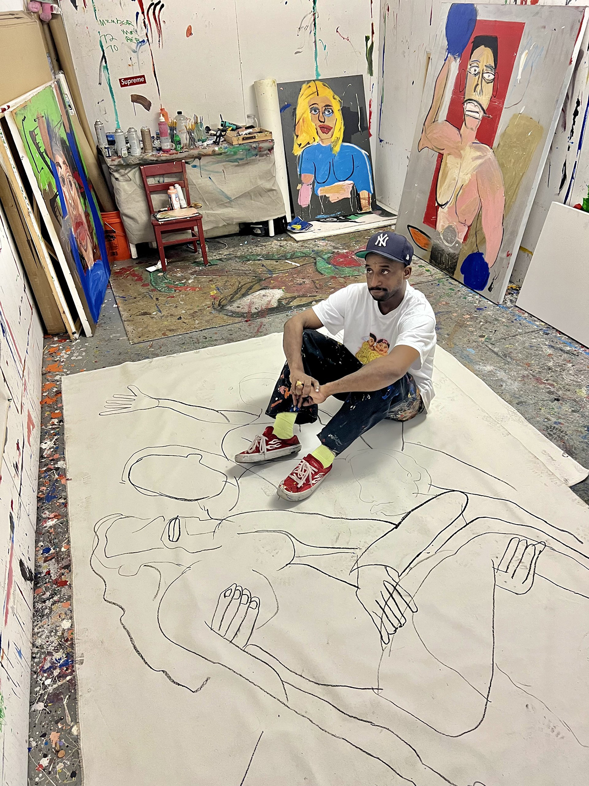A man wearing a white t shirt sitting on an art work on the floor in a studio