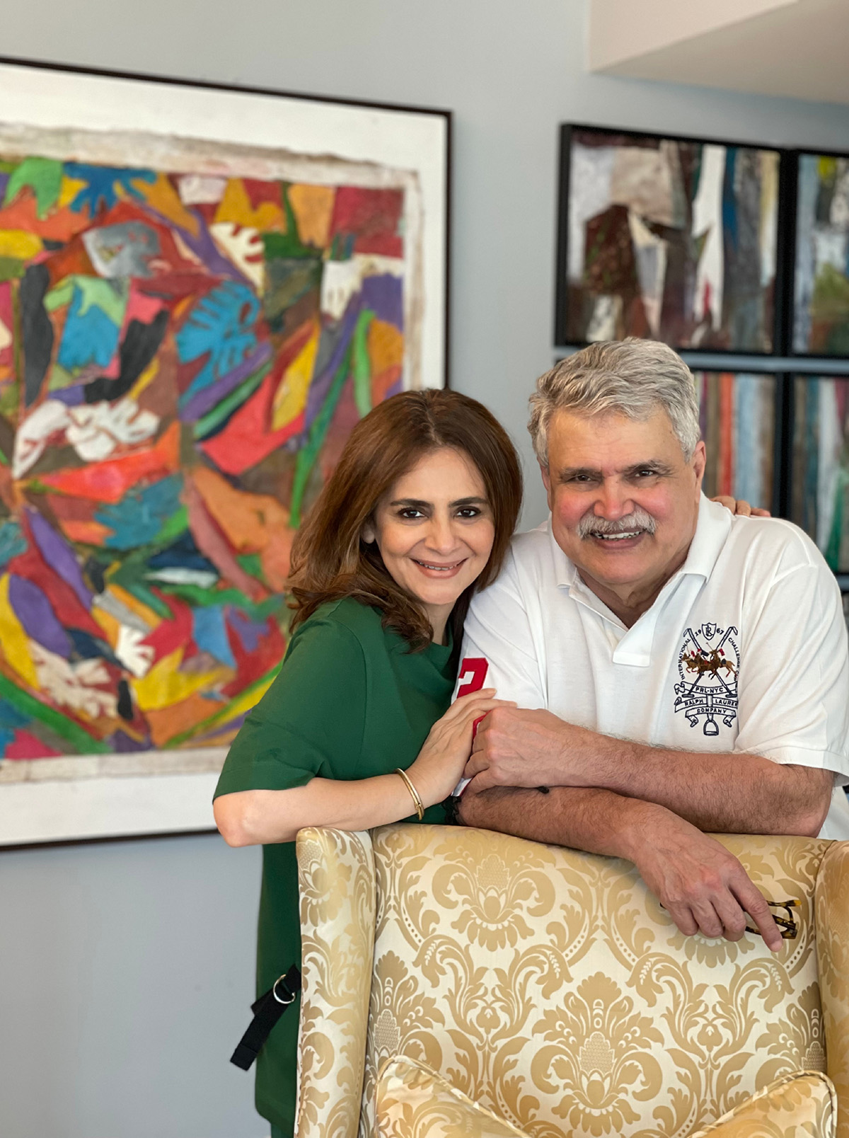 A woman wearing a green top standing next to a pan in a white polo shirt leaning on a gold chair with art behind them on the wall