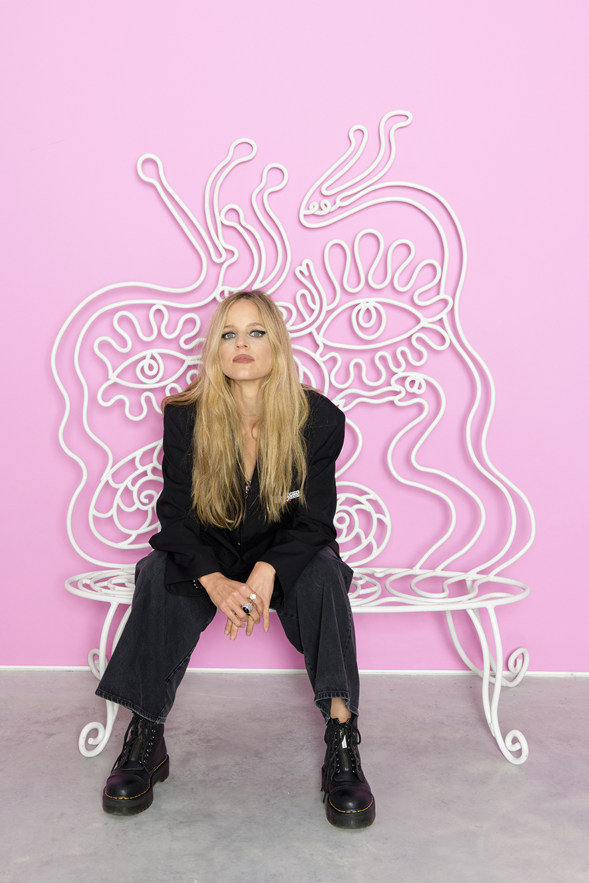 A blonde woman wearing a black suit sitting in a pink room on a white bench with squiggles coming out of it