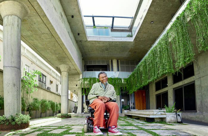 A man sitting in a wheelchair, wearing red trousers, in a courtyard of a building with plants hanging over the interior balconies