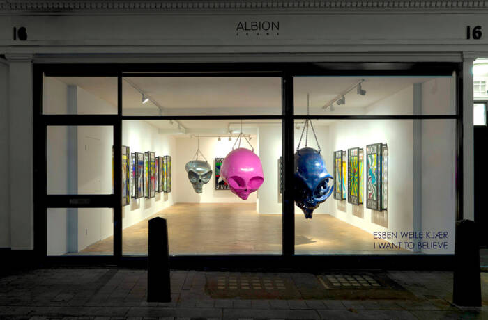 The window of a gallery with hanging coloured giant skulls in the room surrounded by pictures