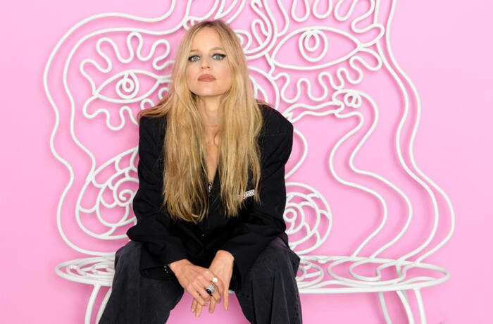 A blonde woman wearing a black suit sitting in a pink room on a white bench with squiggles coming out of it