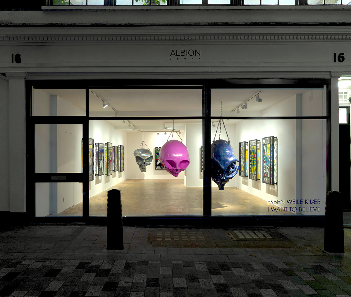 The window of a gallery with hanging coloured giant skulls in the room surrounded by pictures