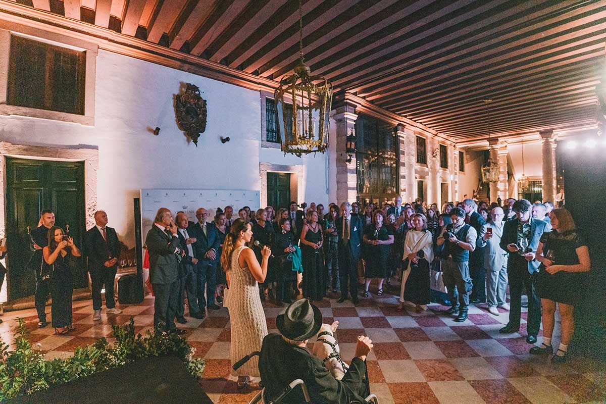 A room full of people and a woman giving a speech with a man in a wheelchair holding a dog in his lap