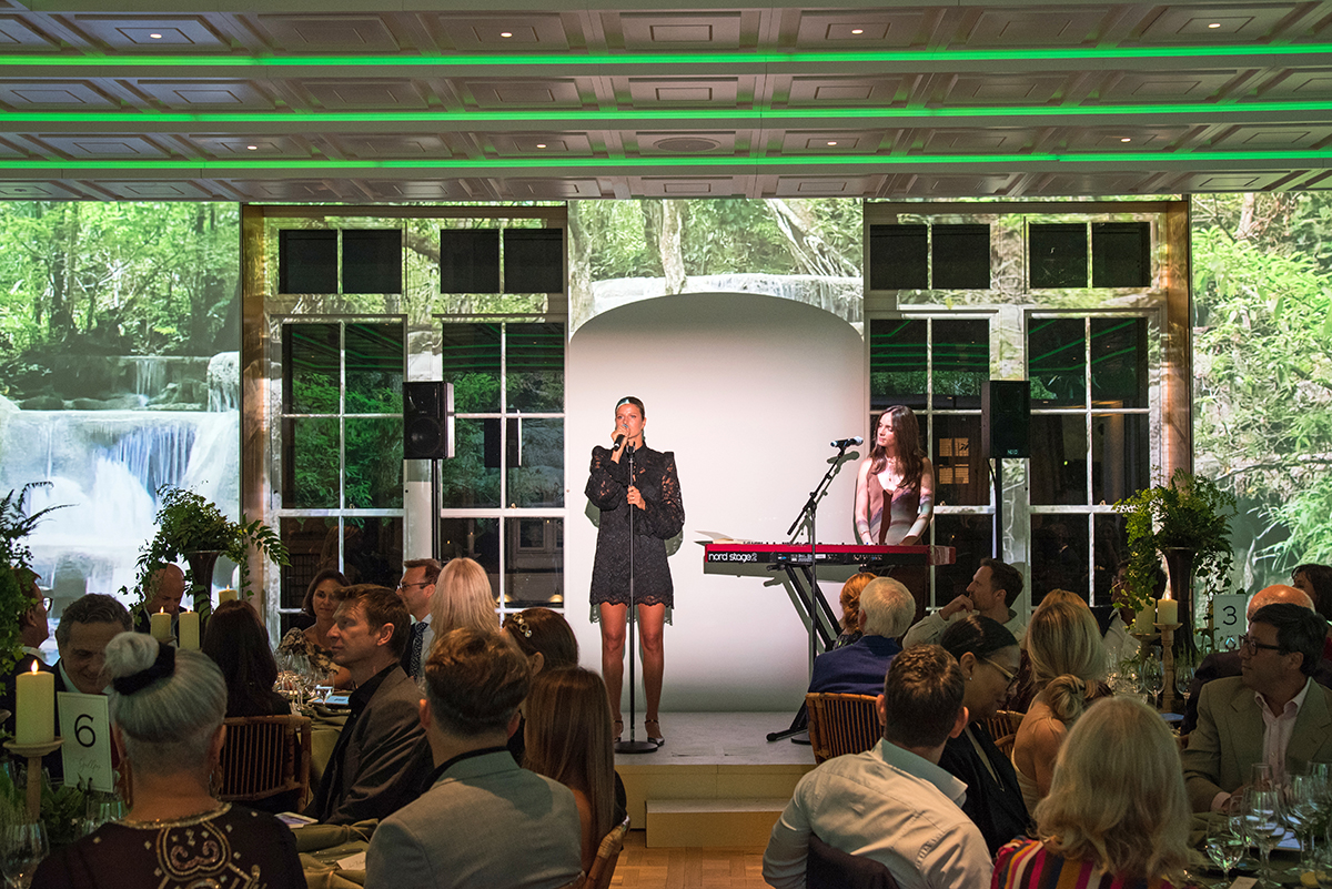 A woman singing on stage next to a person on a keyboard with green lights above her and people watching sitting at tables