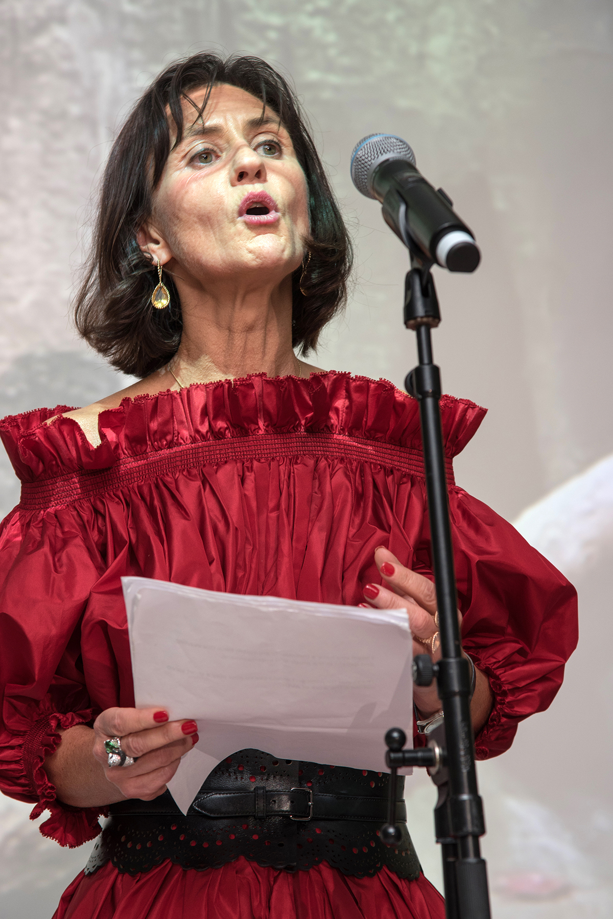 A woman with short black hair wearing a red puffy off the shoulder dress giving a speech