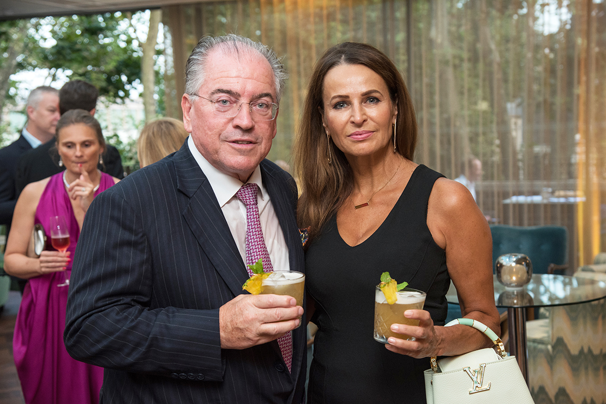 A man in a suit holding a drink next to a woman wearing black dress also holding a drink