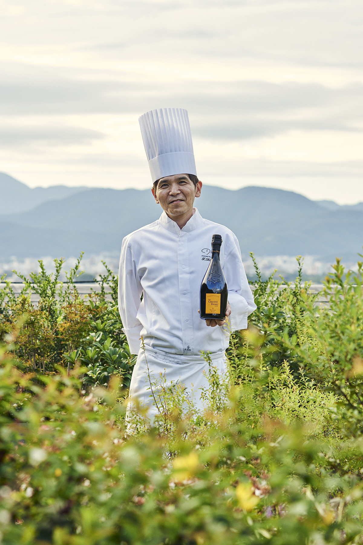 A chef wearing a long white hat and uniform holding a bottle of champagne in a field