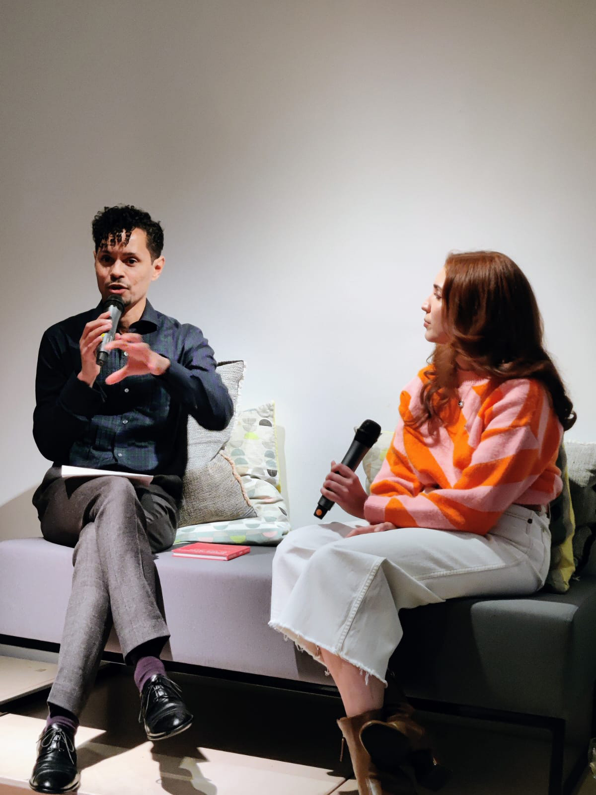 A woman wearing an orange and pink top speaking to a man sitting on a couch
