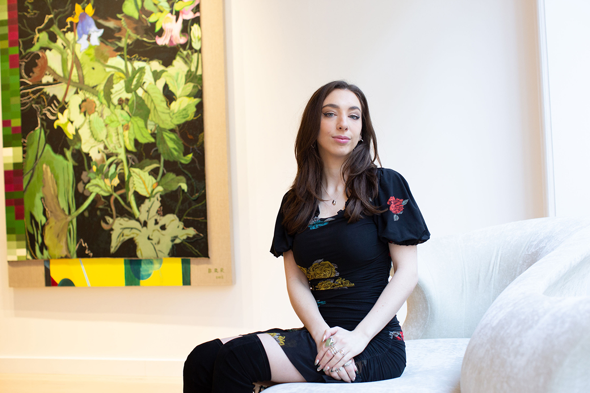 A girl with dark hair wearing a black dress and black boots sitting on a chair with a green painting behind her