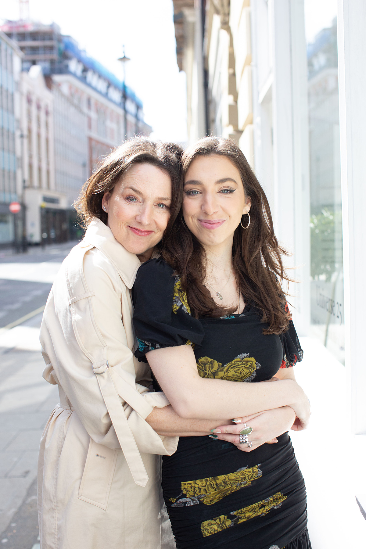 A mother wearing a trench coat hugging her daughter who is wearing a black short sleeve dress