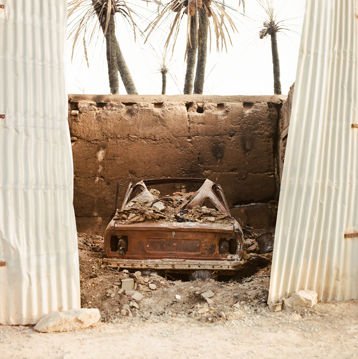 A burned and deserted car in the sand by a wall