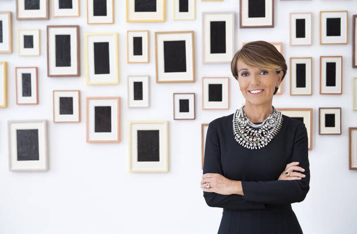 A woman wearing a balck top and large diamond necklace standing net to a wall with frames and black boxes in the frames