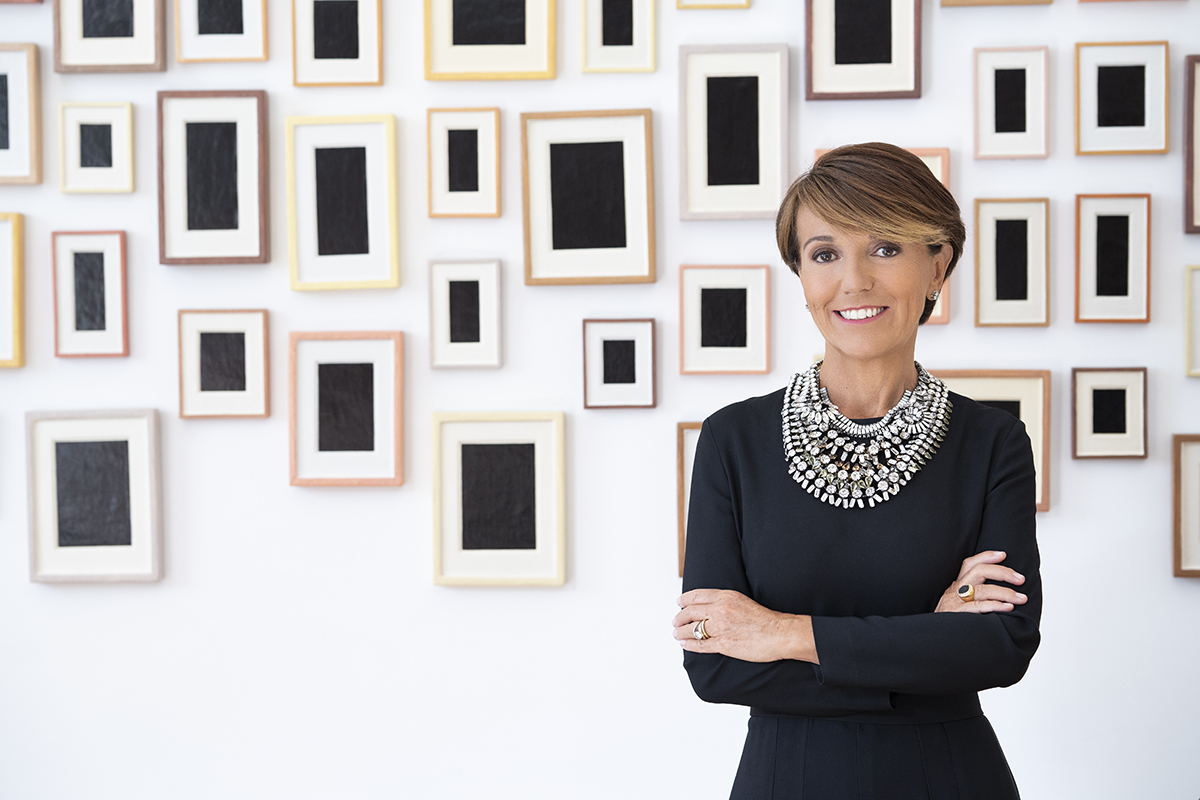 A woman wearing a balck top and large diamond necklace standing net to a wall with frames and black boxes in the frames
