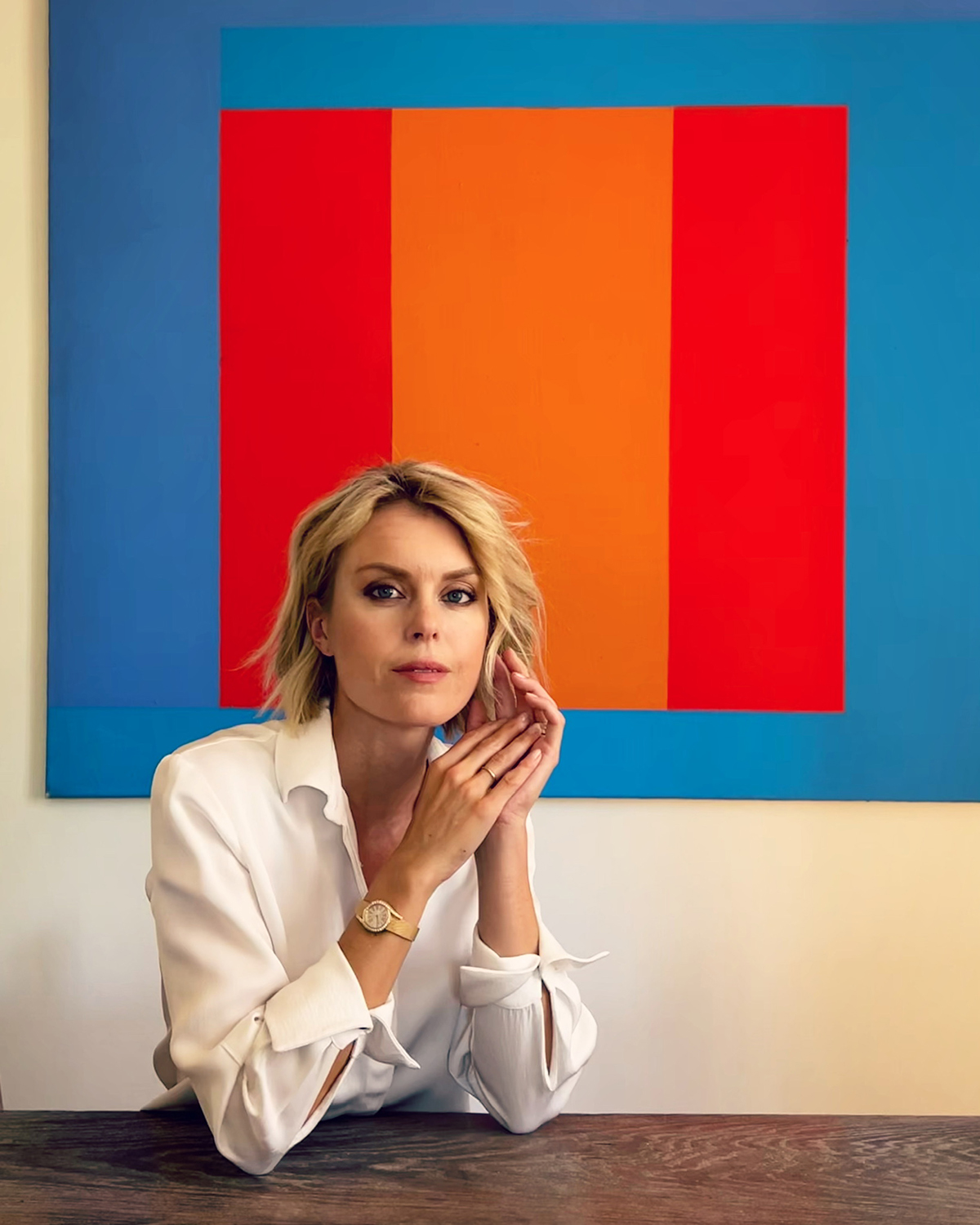 A blonde woman wearing a white shirt sitting in front of a blue orange and red block colour painting