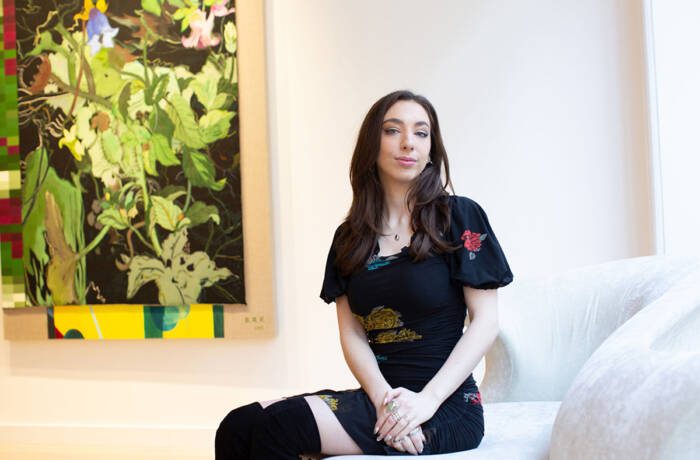 A girl with dark hair wearing a black dress and black boots sitting on a chair with a green painting behind her