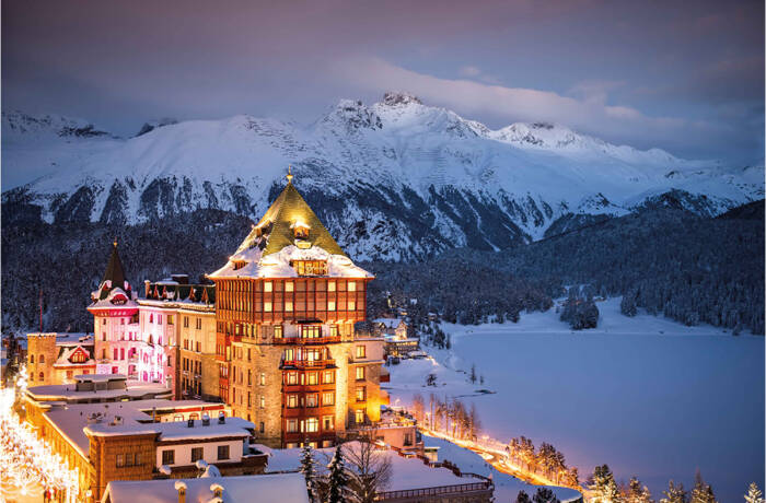 A lit up hotel at night in front of mountains covered in snow