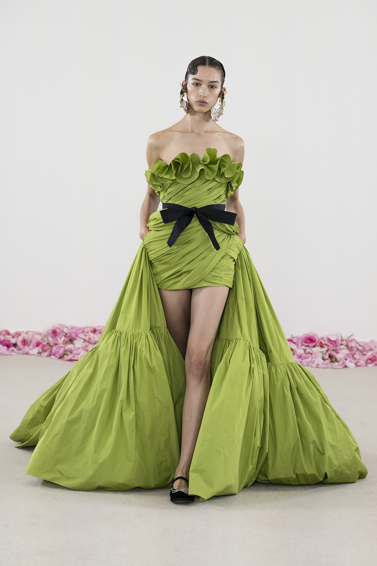 A woman wearing a long green ball gown that is long at the back and short at the front with a black bow around her waist
