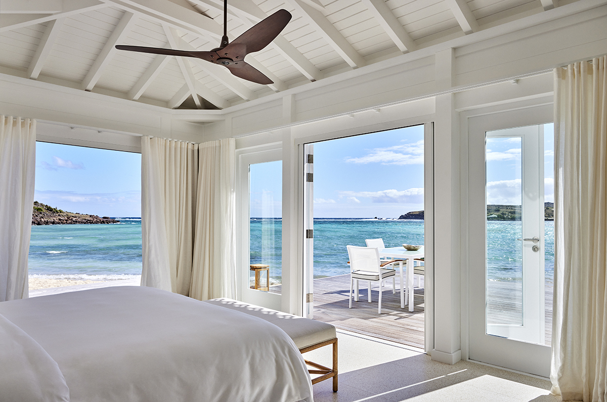 A white bedroom overlooking a beach with turquoise sea and chairs and tables outside the room
