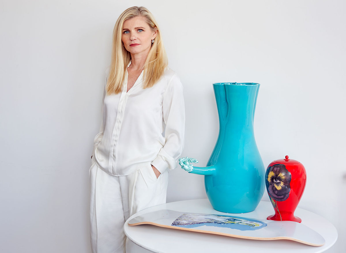 A blonde woman wearing a white shirt and white trousers standing next to a table with a blue vase and a red ornament