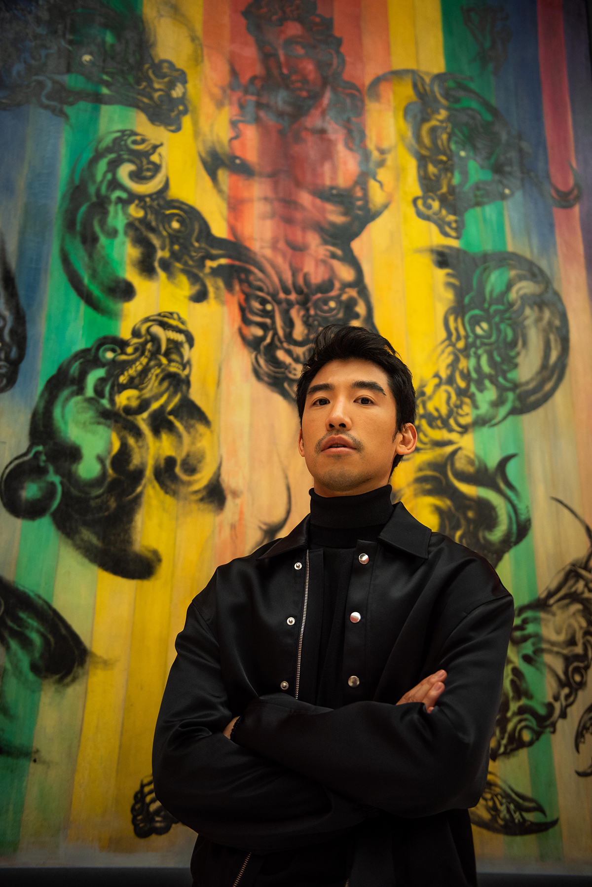 A man wearing black with his arms folded standing in front of a red, black, green and yellow asian painting