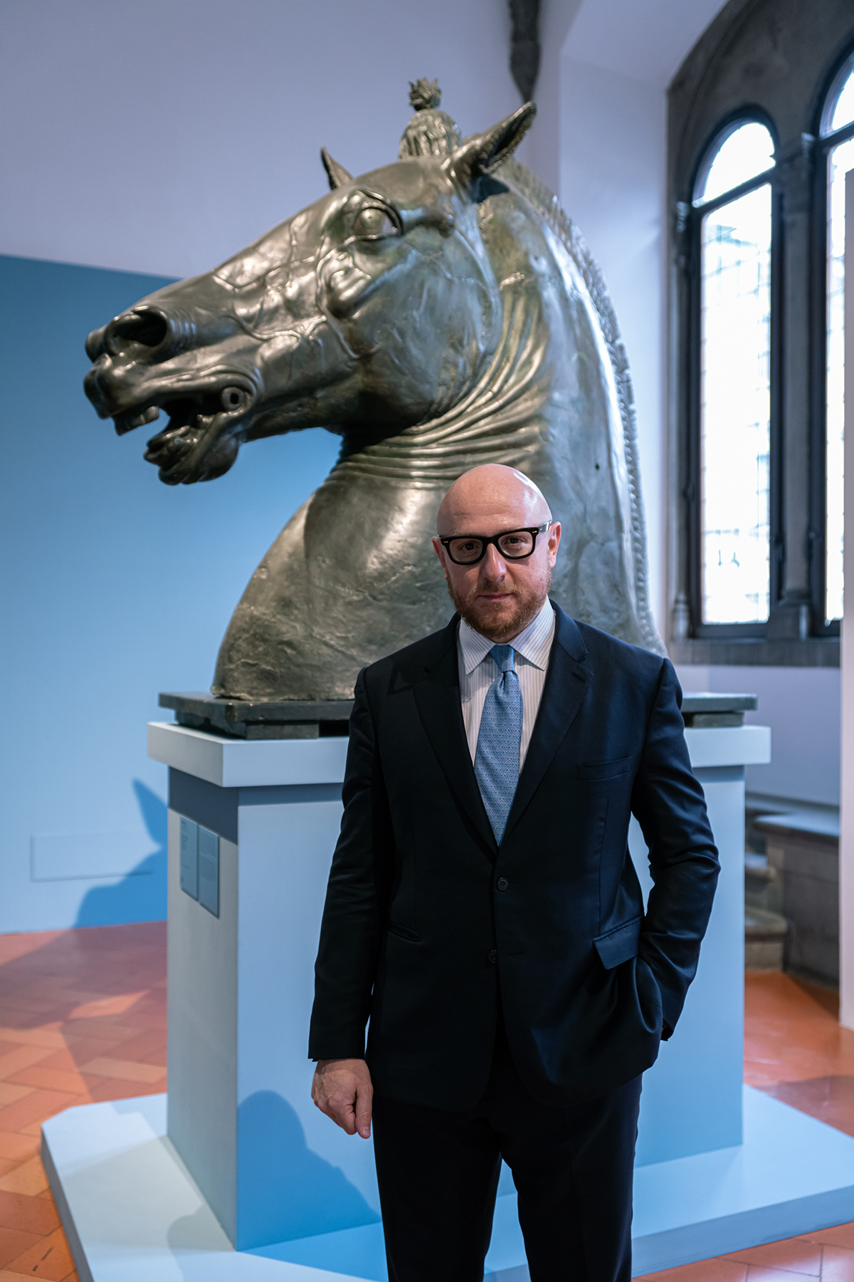 A man wearing a suit and blue tie standing in front of a bust of a horse