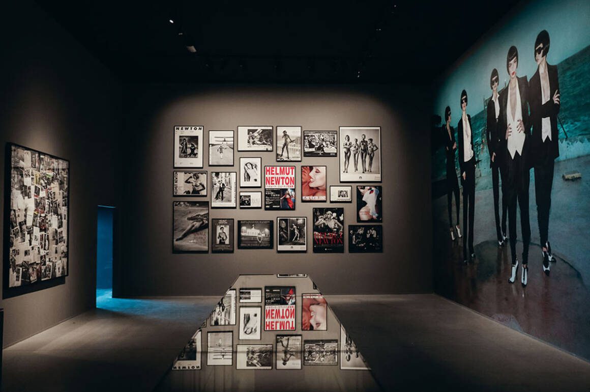 photographs hanging on a wall with Helmut Newton in white a ona. red background witten on a wall and a projected image on an adjacent wall