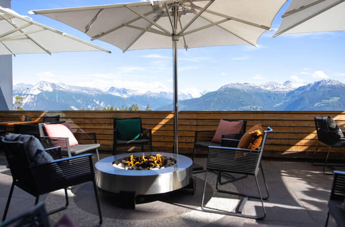 A terrace with a fire pit in the middle surrounded by chairs with cushions and a parasol